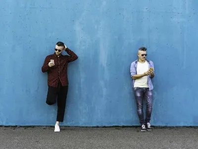 Cool men looking at mobile phone lean against blue wall outdoors - Couple of friends addicted to technology and social networks - Concept of tech, social network and social relationships