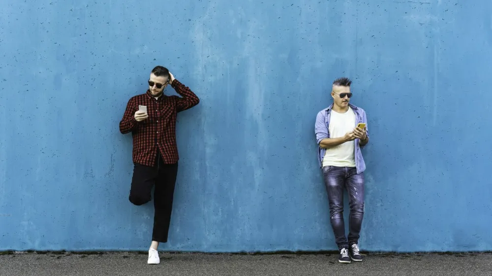 Cool men looking at mobile phone lean against blue wall outdoors - Couple of friends addicted to technology and social networks - Concept of tech, social network and social relationships