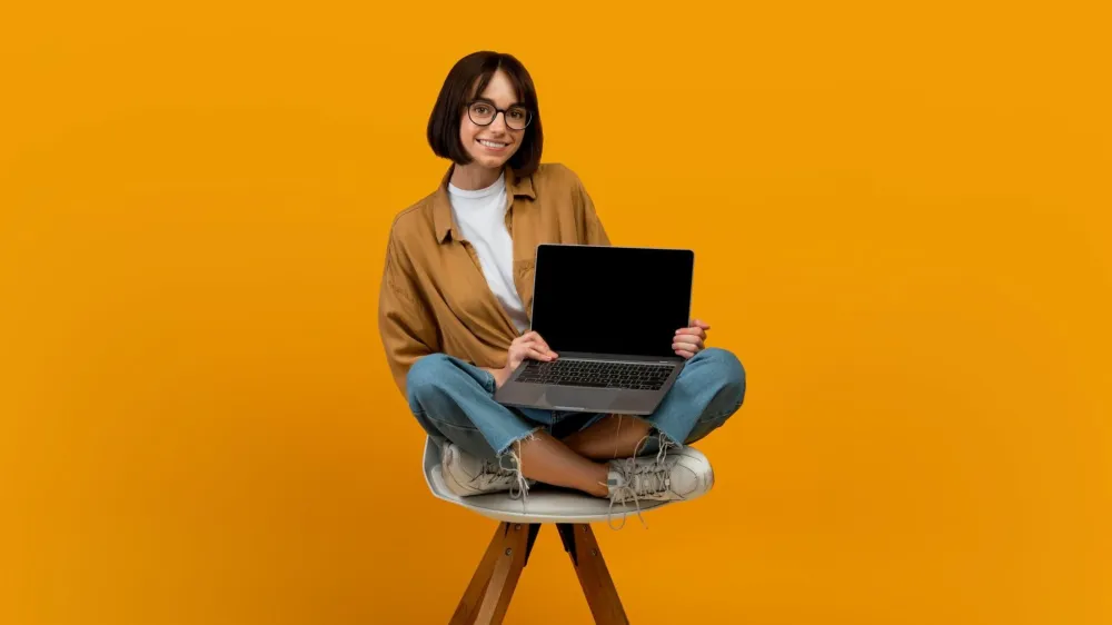 Website advertisement. Happy young lady with laptop showing empty computer screen recommending online service, sitting in chair over yellow wall background. Mockup