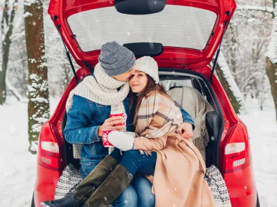 Couple in love sitting in car trunk drinking hot tea in snowy winter forest and chatting. People relaxing outdoors during road trip