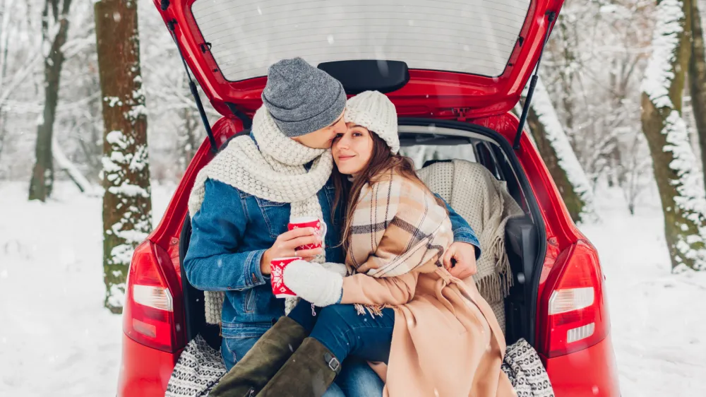 Couple in love sitting in car trunk drinking hot tea in snowy winter forest and chatting. People relaxing outdoors during road trip