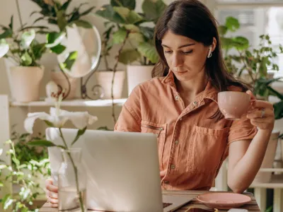 Young dark-haired mixed race woman using a laptop,drinking coffee at home.Cozy home interior with indoor plants.Remote work, business,freelance,online shopping,e-learning,urban jungle concept.