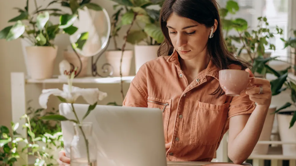 Young dark-haired mixed race woman using a laptop,drinking coffee at home.Cozy home interior with indoor plants.Remote work, business,freelance,online shopping,e-learning,urban jungle concept.