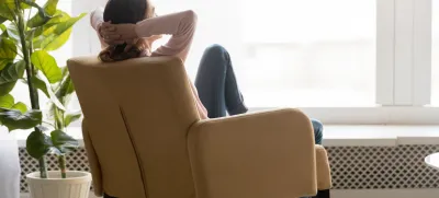 Back view of young happy woman sitting in comfortable armchair, facing shiny window, crossing hands behind head, having rest after housework, resting on weekend, lazy day concept.