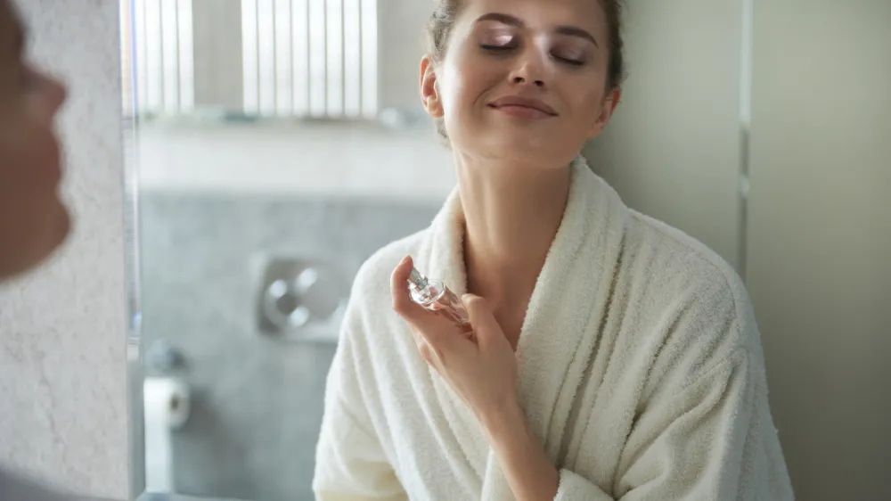Self care concept. Waist up reflection of young smiling woman in white bathrobe enjoying her morning while applying favorite perfume before bathroom mirror