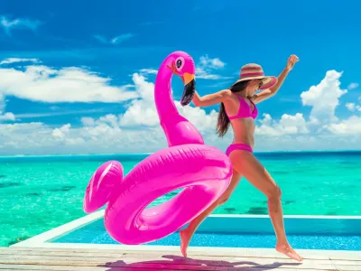 Vacation fun woman in bikini with funny inflatable pink flamingo pool float running of joy jumping by infinity swimming pool. Girl enjoying travel holidays at resort luxury overwater bungalow travel.