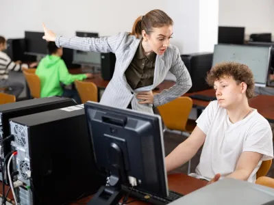 Strict young female teacher reprimanding negligent teenage student sitting at computer in classroom during lesson