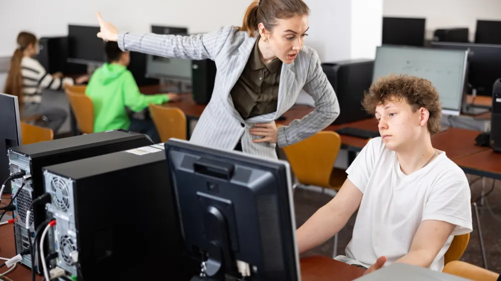 Strict young female teacher reprimanding negligent teenage student sitting at computer in classroom during lesson