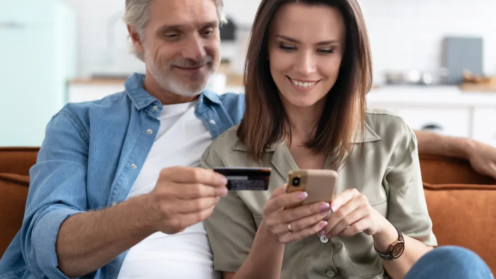 Smiling caucasian couple involved in online shopping, entering payment information from credit bank card in mobile application, satisfied with quick money transfer, modern tech concept.