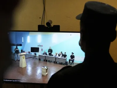 An officer stands in front of a TV screen showing Russian opposition leader Alexei Navalny, standing, speaking between his lawyers in a courtroom, via video link provided by the Russian Federal Penitentiary Service, during a preliminary hearing in the colony, in Melekhovo, Vladimir region, about 260 kilometers (163 miles) northeast of Moscow, Russia, on Monday, June 19, 2023. A Russian court has opened a new trial of imprisoned Russian opposition leader Alexei Navalny that could keep him behind bars for decades. (AP Photo/Alexander Zemlianichenko)