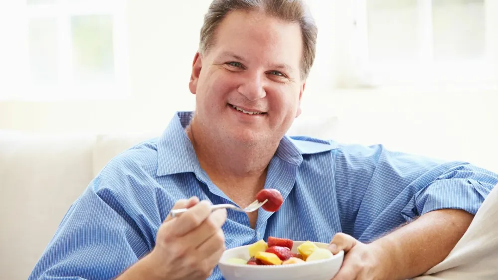 Overweight Man Sitting On Sofa Eating Bowl Of Fresh Fruit At Home Smiling To Camera