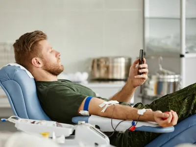 Side view portrait of military man giving blood while laying in chair at blood donation center and watching videos at smartphone
