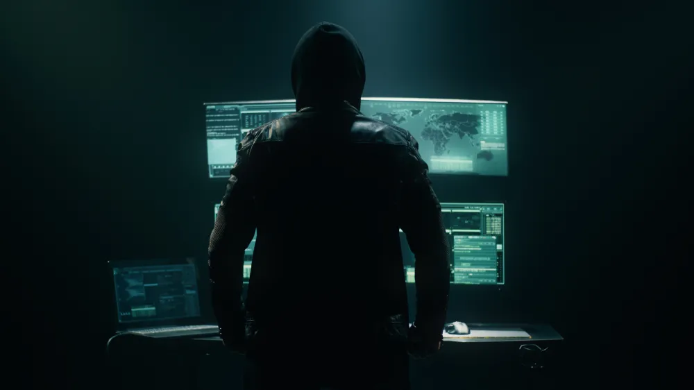 Back view of unrecognizable man in hoodie standing near desk and reading stolen data from computer, monitors in dark room before massive cyber attack on servers.