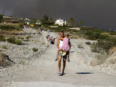 A man carries a child as they leave an area where a forest fire burns, on the island of Rhodes, Greece, Saturday, July 22, 2023. A large wildfire burning on the Greek island of Rhodes for a fifth day has forced authorities to order an evacuation of four locations, including two seaside resorts. (Lefteris Diamanidis/InTime News via AP)