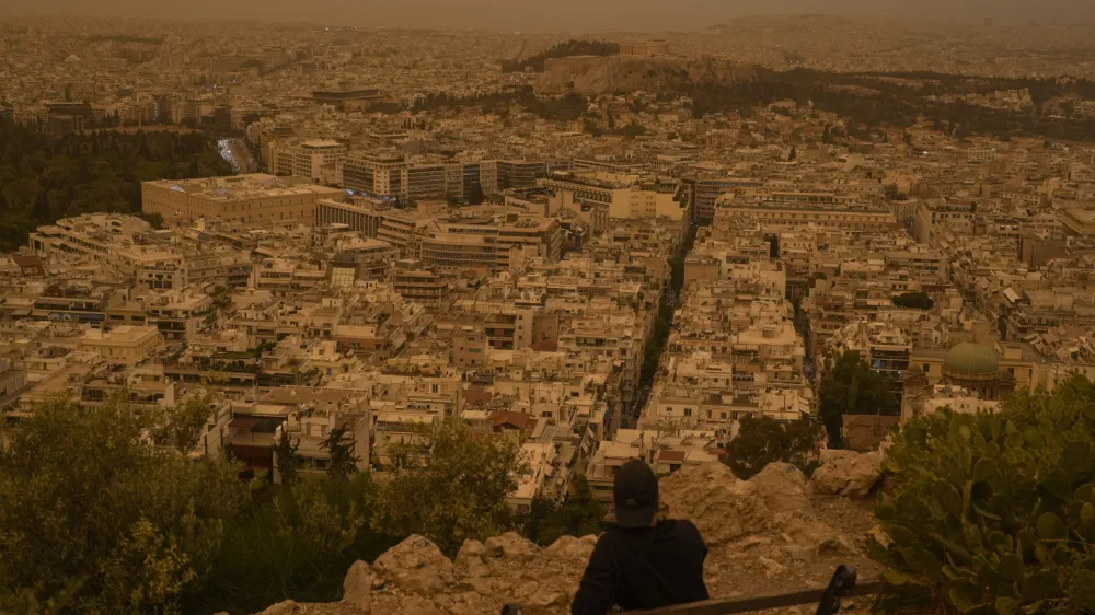 A tourist sits on a bench at the Lycabettus hill as the city of Athens with the ancient Acropolis hill is seen at the background, on Tuesday, April 23, 2024. The Acropolis and other Athens landmarks took on Martian hues Tuesday as stifling dust clouds blown across the Mediterranean Sea from North Africa engulfed the Greek capital. (AP Photo/Petros Giannakouris)