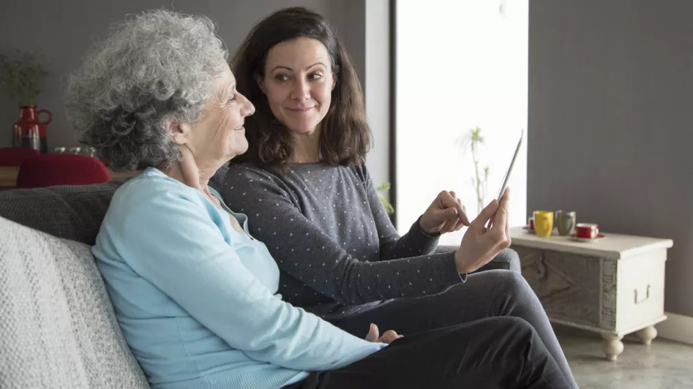 Smiling senior woman and her daughter using tablet computer. Mother and daughter sitting on couch with home interior in background. Technology concept. / Foto: Mangostar_studio