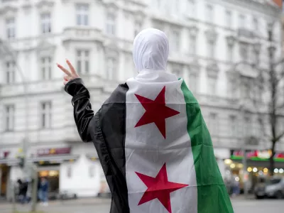 A Syrian girl flashes victory sign in a street in Berlin while wearing a Syrian opposition flag, Tuesday, Dec. 10, 2024. (AP Photo/Ebrahim Noroozi)