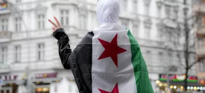 A Syrian girl flashes victory sign in a street in Berlin while wearing a Syrian opposition flag, Tuesday, Dec. 10, 2024. (AP Photo/Ebrahim Noroozi)