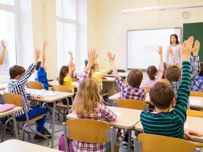 education, elementary school, learning and people concept - group of school kids with teacher sitting in classroom and raising hands