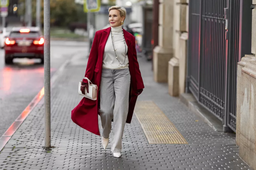 Stylish Woman in Burgundy Coat Walks City Street. Wearing Fashionable clothing White Turtleneck Sweater, Pearl Necklace and Grey Jeans, Carrying White Bag. Confident and Chic Mature Fashion Model