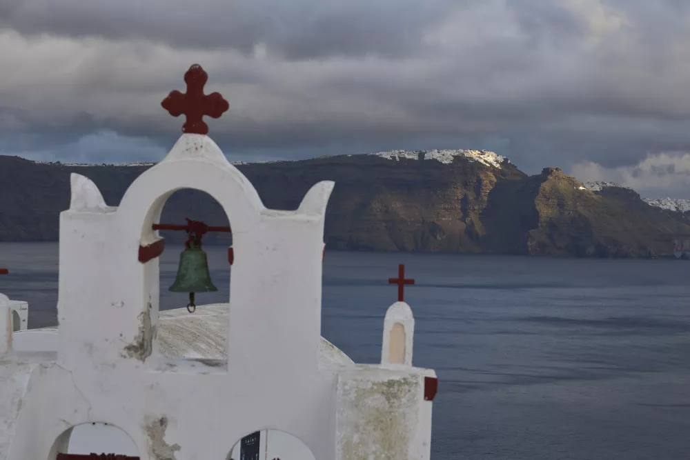 A bell towel of an Orthodox church in the town of Oia on the earthquake-struck island of Santorini, Greece, as the main town of Fira is litted by the sun in the background on Tuesday, Feb. 4, 2025. (AP Photo/Petros Giannakouris)