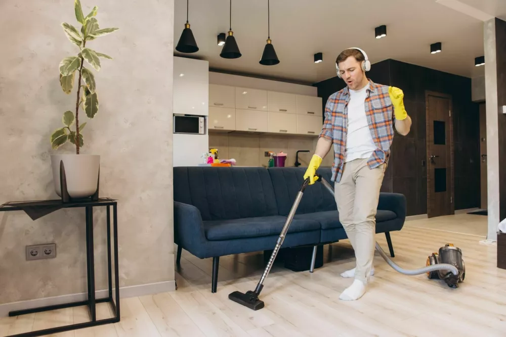 Young man is vacuuming the floor in living room and dancing while listening to music with headphones, enjoying cleaning his apartment / Foto: Anatoliycherkas