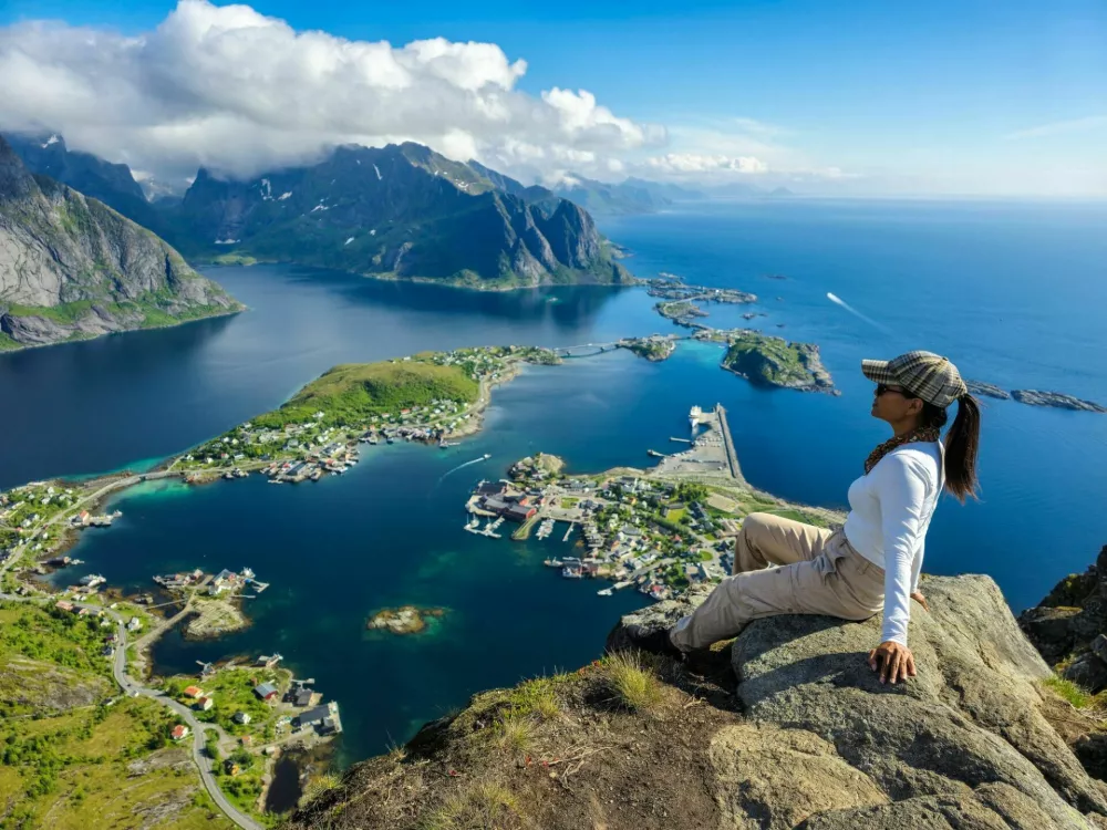 An adventurer sits atop a rocky ledge, soaking in the panoramic beauty of the Lofoten Islands as the sun rises, illuminating the tranquil waters and rugged mountains. Reinebringen Lofoten Norway / Foto: Fokkebok