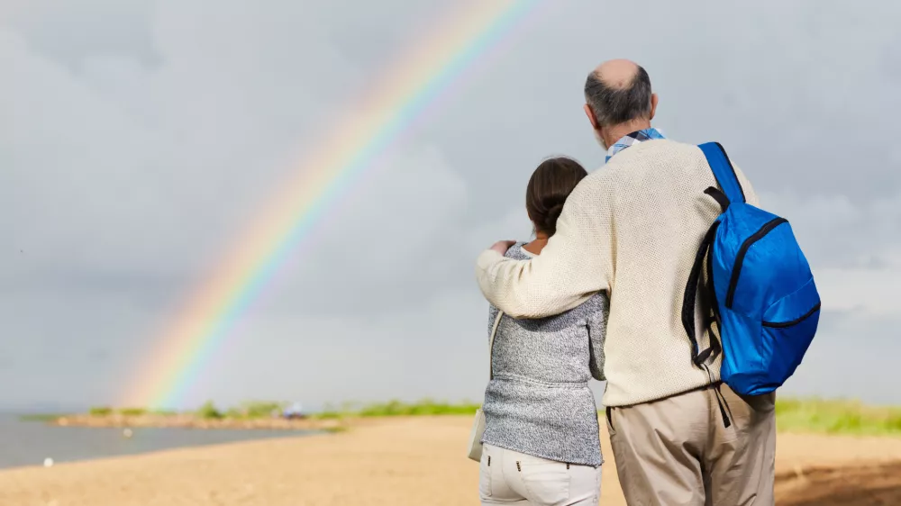 Affectionate aged couple looking at rainbow against grey cloudy sky / Foto: Shironosov