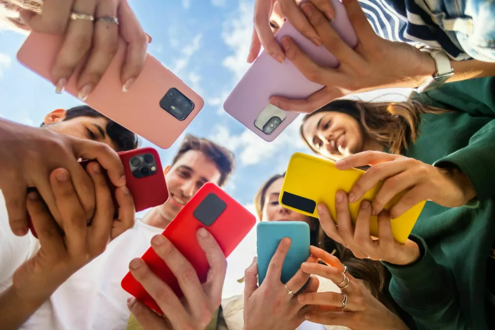 Young group of people using mobile phone device standing together in circle outdoors. Millennial friends addicted to social media app, betting or playing video game on platform online. / Foto: Xavier Lorenzo