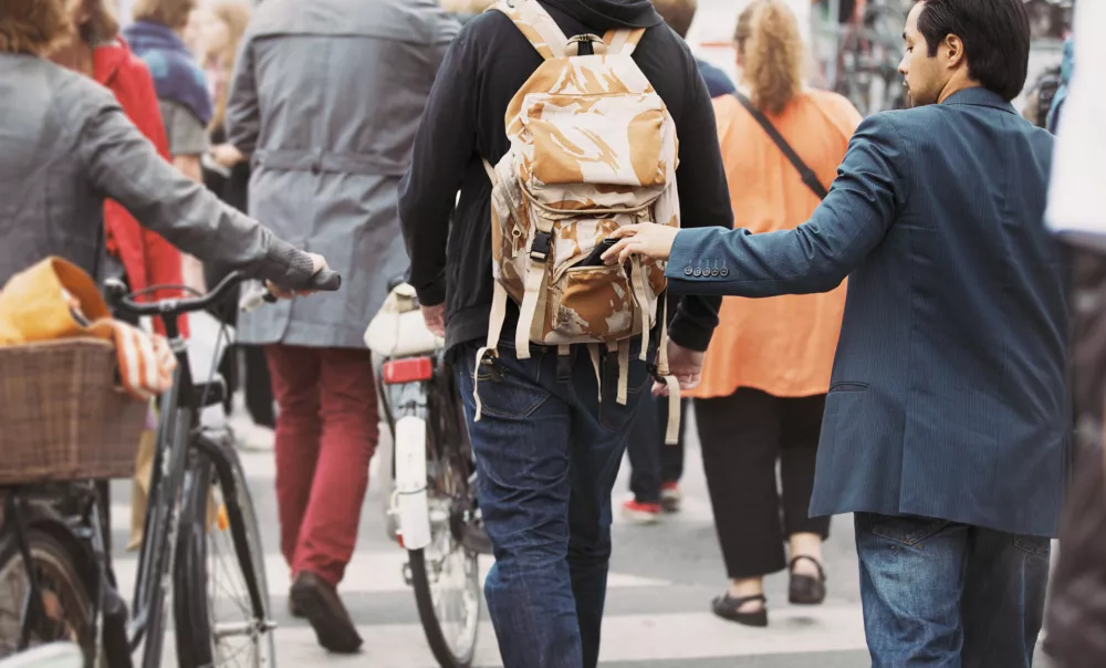Young man taking wallet from backpack of a man walking on street during daytime. Pickpocketing on the street during daytime