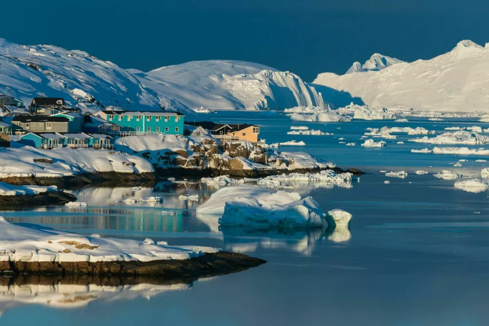 Colorful houses line the shores of a serene bay in the Arctic, surrounded by icebergs and snow-covered mountains, reflecting the warm glow of the sunset on the water.