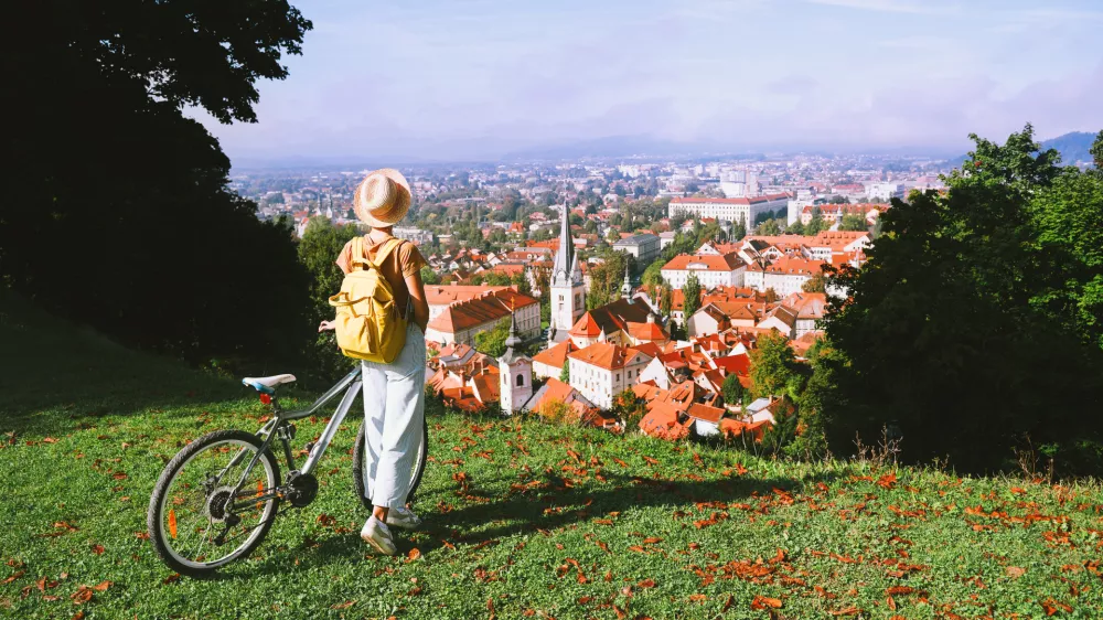 Travel Slovenia Europe. Woman tourist looking at panoramic view of cityscape with red roofs of Ljubljana from City Castle. Young girl with bicycle on top of green hill explores sights and local living / Foto: Nataliaderiabina