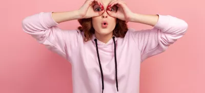 Portrait of amazed teen girl with curly hair in hoodie looking through binoculars gesture and expressing surprise, zooming vision, exploring distance. Indoor studio shot isolated on pink background / Foto: Khosrork