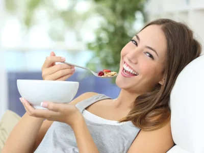﻿Happy girl eating cereals with fruits from a bowl sitting on a couch at home