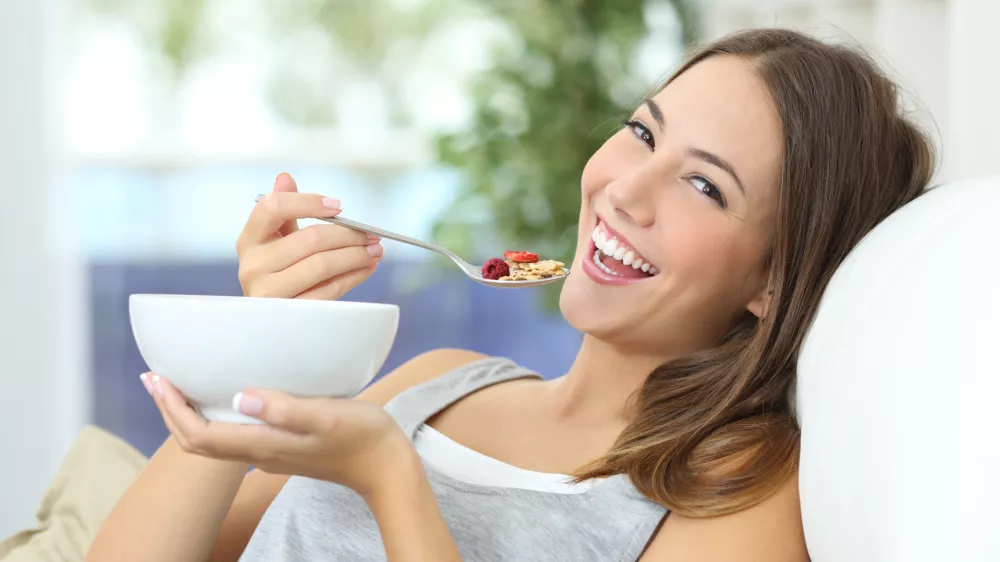 ﻿Happy girl eating cereals with fruits from a bowl sitting on a couch at home