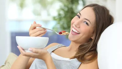 ﻿Happy girl eating cereals with fruits from a bowl sitting on a couch at home