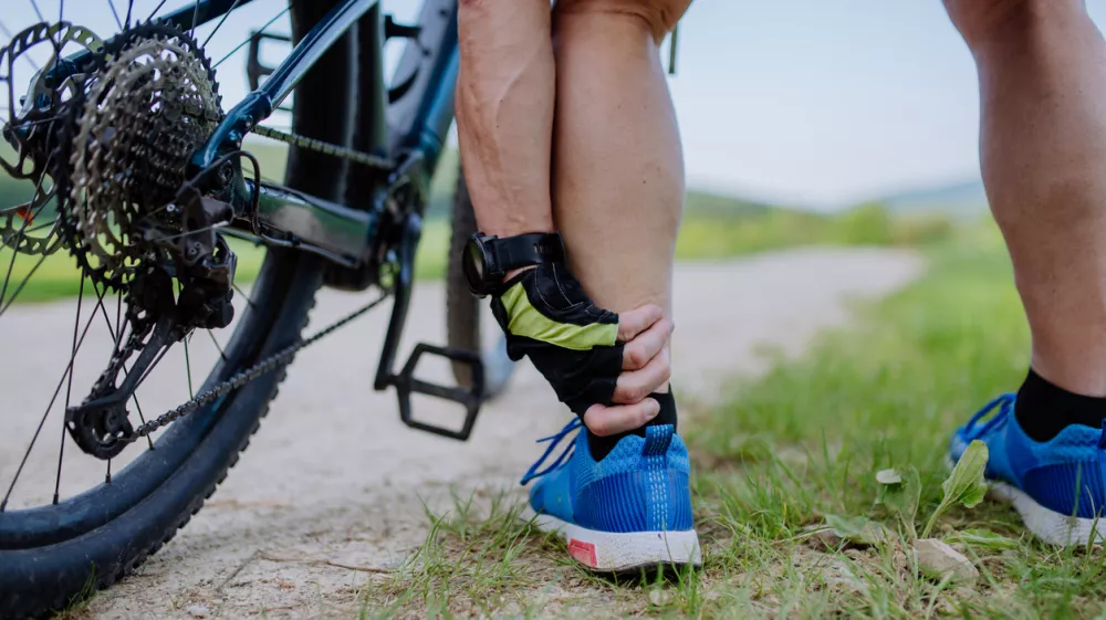 A close-up of active senior man in sportswear suffiering from pain in his ankle after cycling, in park in summer.