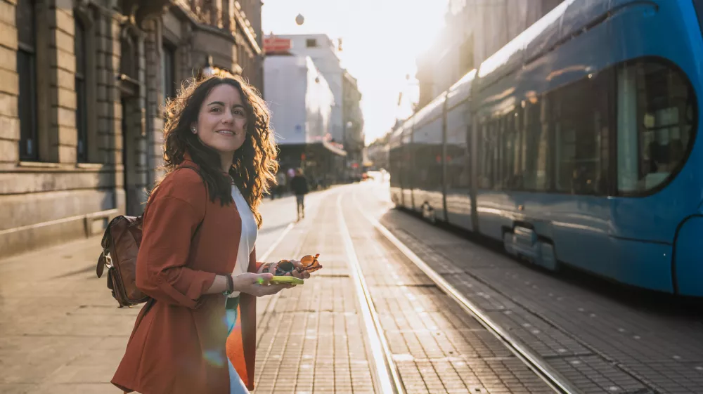 Woman with phone and sunglasses walking across city street next to blue tram. Concept: urban lifestyle, travel experience, exploring Croatia.
