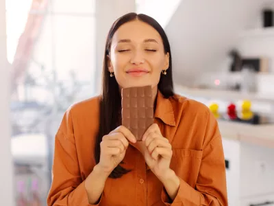 Portrait of happy young woman smelling chocolate bar with closed eyes, want to eat dessert, but on diet, lady sitting in kitchen interior / Foto: Olena Miroshnichenko