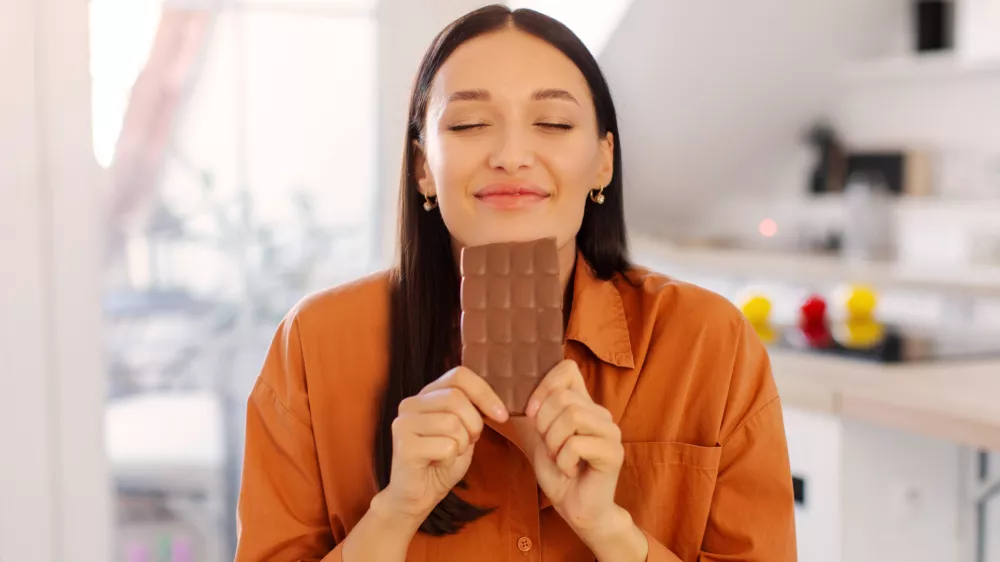Portrait of happy young woman smelling chocolate bar with closed eyes, want to eat dessert, but on diet, lady sitting in kitchen interior / Foto: Olena Miroshnichenko