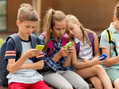 ﻿primary education, friendship, childhood, technology and people concept - group of happy elementary school students with smartphones and backpacks sitting on bench outdoors
