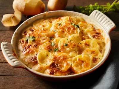 High angle of scalloped gratin potatoes topped with grated cheese and herbs in ceramic baking dish on table with towel and branches of greens / Foto: Foodandstyle, Getty