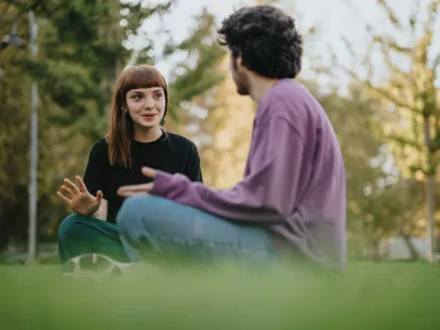 Two young people engage in a friendly conversation while sitting on the grass outdoors. The scene reflects friendship, connection, and relaxation in a green, sunlit environment. / Foto: Qunicastudio