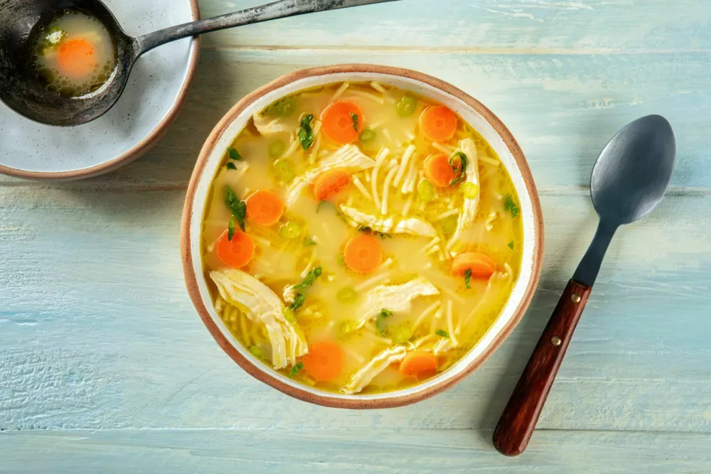 Chicken noodle soup with vegetables, a bowl of healthy stock with a ladle, top shot on a rustic wooden table / Foto: Cook Shoots Food