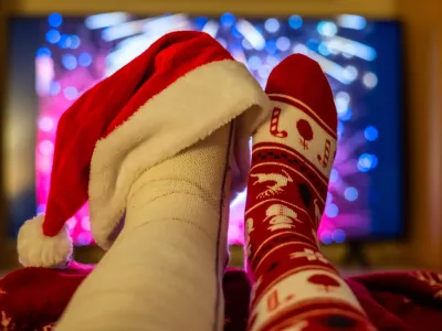 Watching colorful fireworks on tv while relaxing at home, feet up and adorned with a festive christmas sock and a santa hat on a broken foot, celebrating the new year with humor and warmth / Foto: Nanci Santos, Getty Images