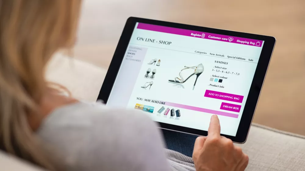 ﻿Closeup of woman doing online shopping on digital tablet at home. Rear view of woman hand touching screen while selecting shoes on ecommerce portal. Lady use e-commerce webshop to buy shoes.