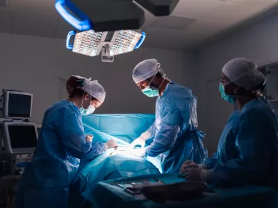 An intricate surgical procedure in progress with healthcare professionals in full surgical gear, under bright operating room lights, emphasizing the modern medical equipment and organization. / Foto: Zamrznutitonovi