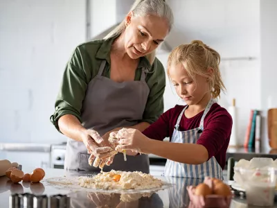 Cute girl learning preparing dough with mature grandmother in kitchen. Lovely old grandma and granddaughter preparing cookies with flour and eggs on kitchen counter. Senior granny in apron teaching her child to make cookies or pie. / Foto: Ridofranz