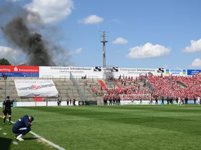 FILED - 11 June 2023, Bavaria, Unterhaching: Unterhaching coach Sandro Wagner (L) sits on the edge of the pitch while thick smoke rises behind the north stand during the the Regionalliga promotion round to the 3rd division soccer match between SpVgg Unterhaching and Energie Cottbus at Sportpark Unterhaching. The Bild paper said the council of the municipality of Unterhaching near Munich has given the green light to sell its 15,000-seat Sportpark Unterhaching stadium to Bayern for ·7.25 million (.5 million). Photo: Angelika Warmuth/dpa