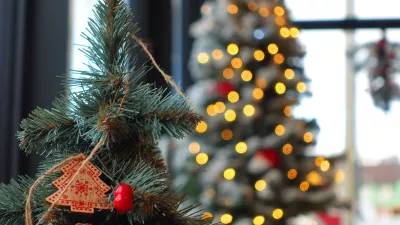 A decorated Christmas tree branch featuring a small wooden tree ornament and bright red berry detail, set against a softly blurred background of warm glowing lights. The bokeh highlights classic seasonal charm, handcrafted holiday style, and cozy winter ambiance. Perfect for festive campaigns, December storytelling, interior Christmas promotion, greeting card visuals, and winter celebration mood boards